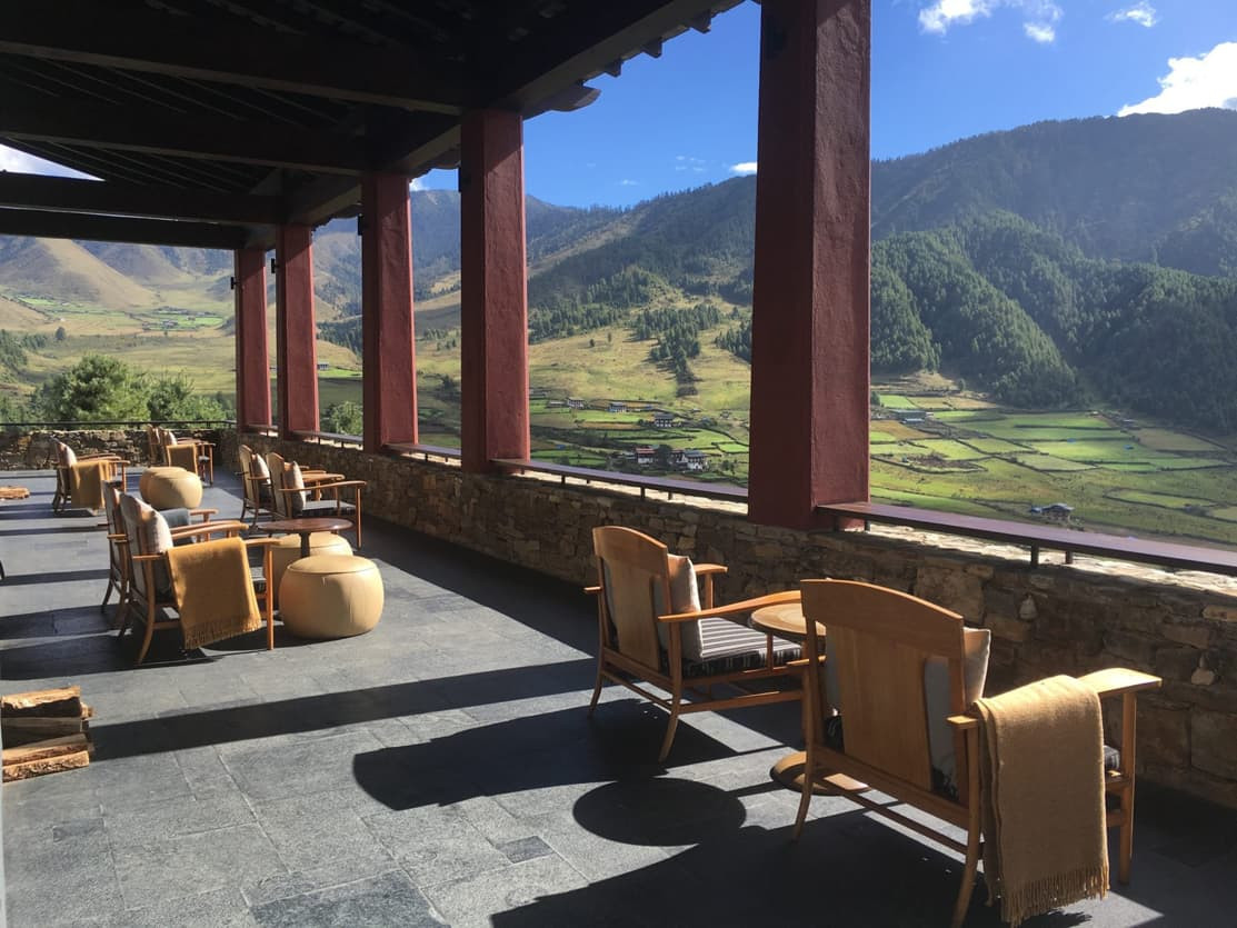 A scenic terrace overlooking lush green valleys and mountains, with wooden chairs and tables arranged on a stone floor. The sunlight casts gentle shadows, enhancing the serene atmosphere.