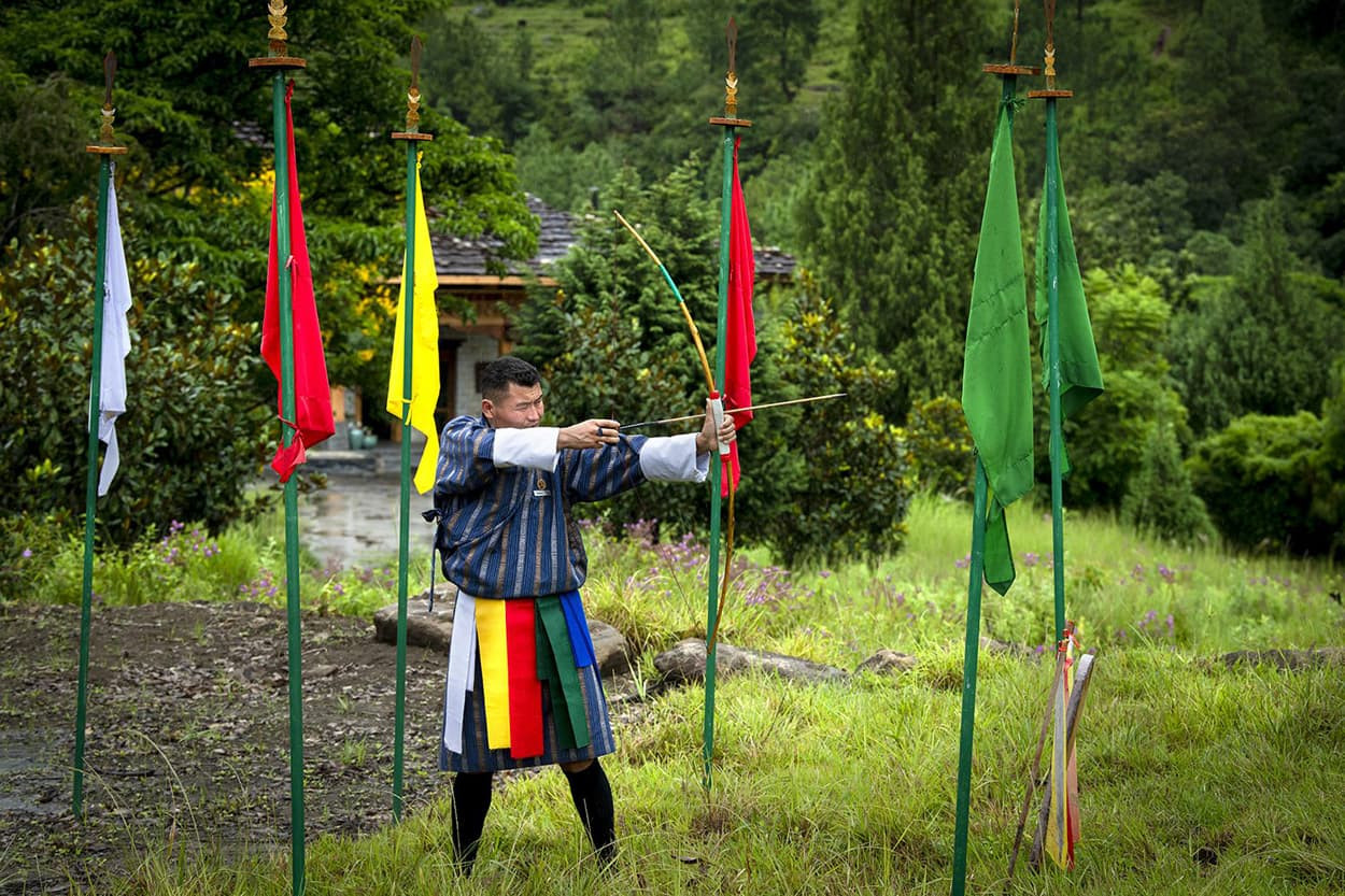 A person in traditional Bhutanese clothing aims an arrow at colored flags set on poles in a grassy area, surrounded by lush greenery.