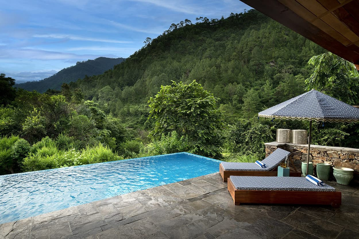 A luxurious infinity pool overlooking lush green hills, with a shaded lounge area featuring two wooden loungers and a stylish umbrella. The serene landscape includes dense trees and a blue sky in the background.