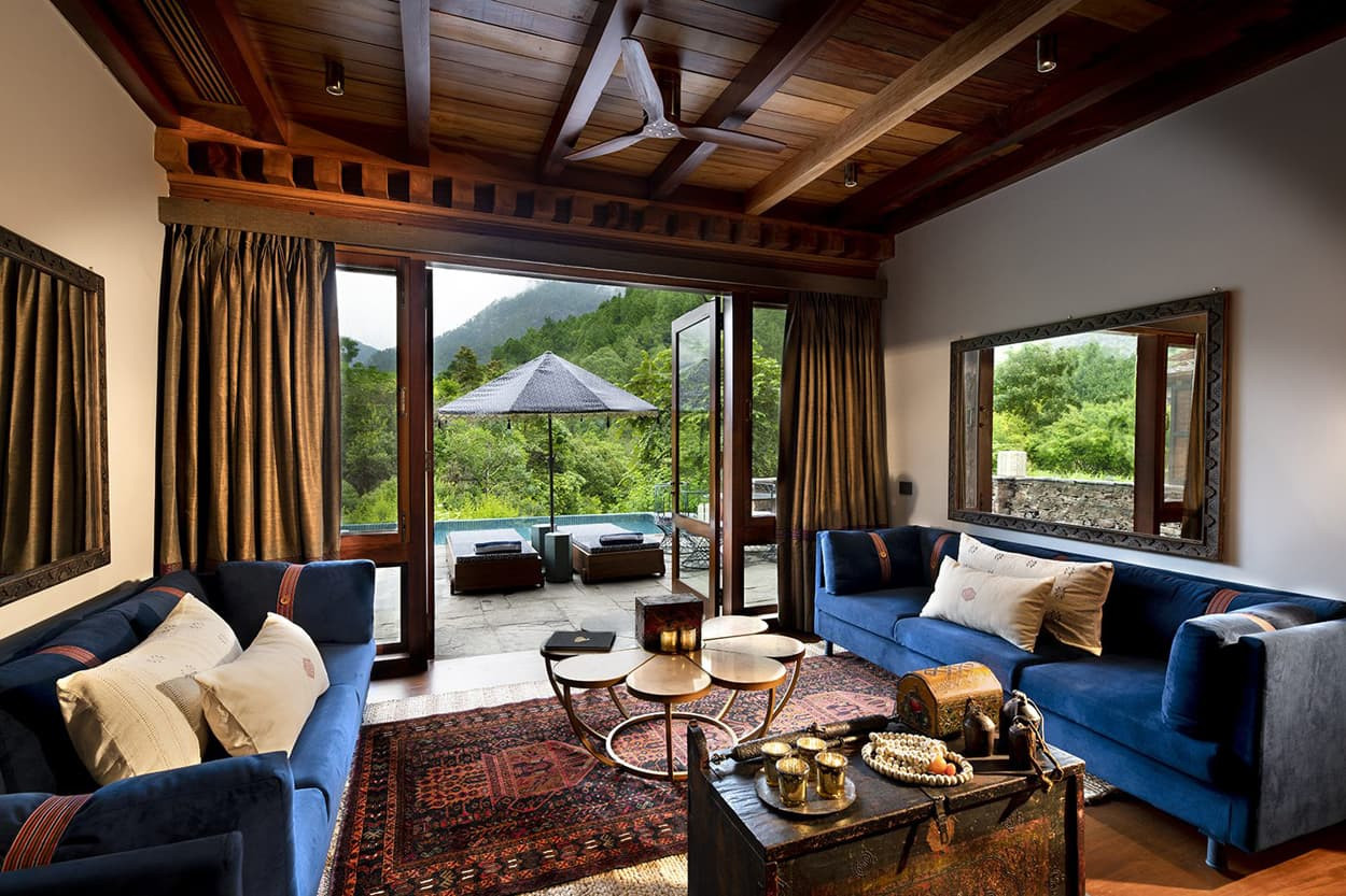Stylish living room with blue sofas, a coffee table, and decorative items. Large windows open to a scenic view of green mountains. Wooden ceiling and warm lighting create a cozy atmosphere.
