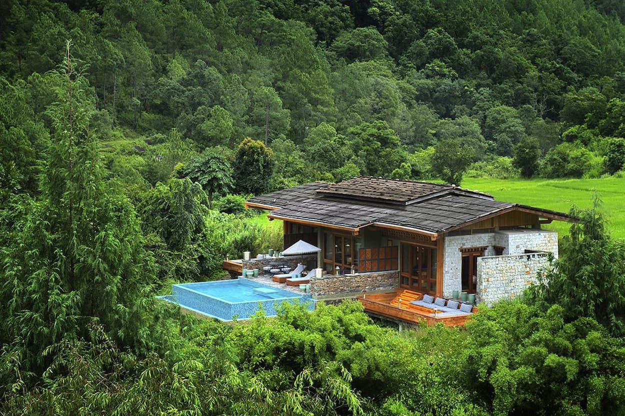A modern villa nestled in lush greenery, featuring a clear blue swimming pool, stone and wooden architecture, and comfortable outdoor seating. The surrounding landscape is filled with dense trees and vibrant vegetation.
