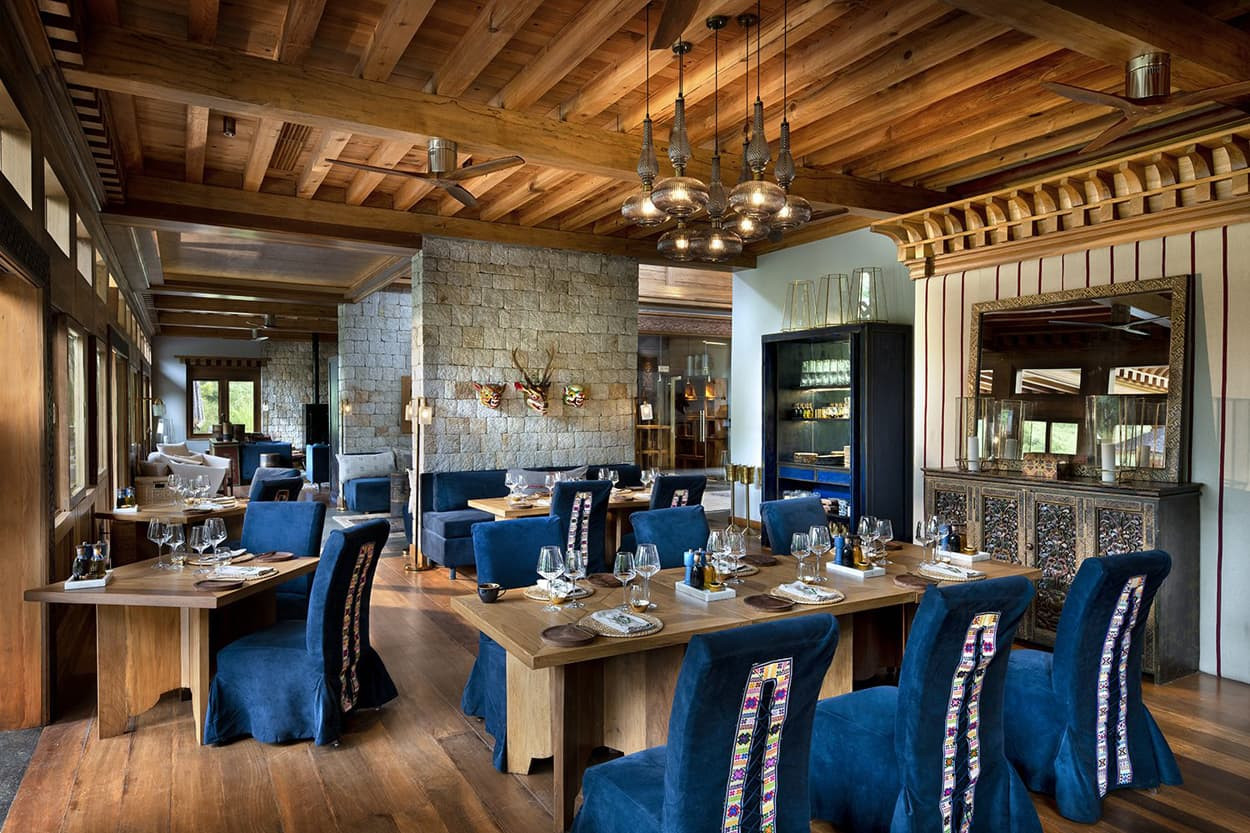 A spacious restaurant interior featuring wooden beams and stone walls. Elegant tables are set with fine dining utensils, surrounded by rich blue upholstered chairs. Ambient lighting from decorative fixtures creates a warm atmosphere.