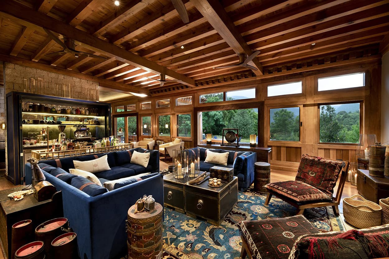 A cozy living room features blue sofas, ornate rugs, and wooden beams. Large windows offer a view of greenery outside. Stylish decor includes a shelf filled with various items and decorative cushions.