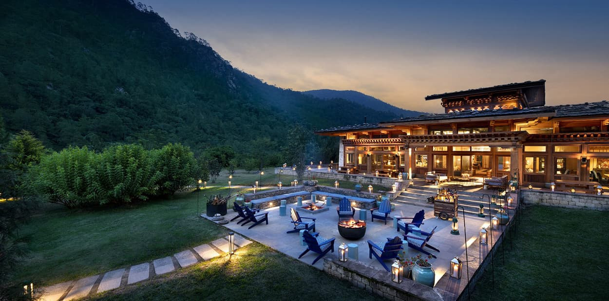 A scenic view of a luxurious wooden house surrounded by mountains at dusk, featuring a spacious outdoor area with fire pits, seating, and lanterns. Lush greenery lines the property, creating a tranquil atmosphere.