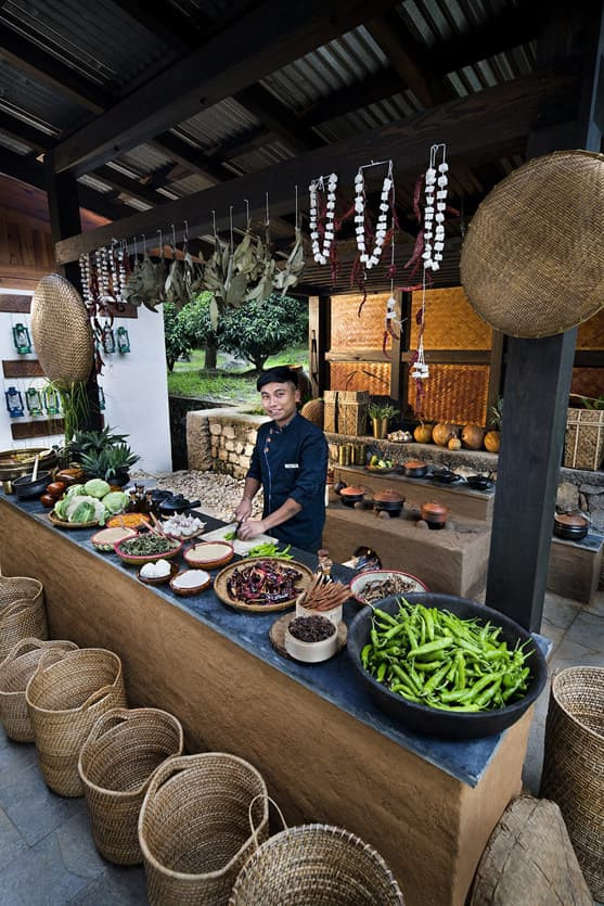 A chef stands behind a rustic outdoor kitchen counter filled with fresh vegetables, spices, and traditional cooking tools. Baskets and dried herbs hang overhead, creating a vibrant, inviting culinary atmosphere.