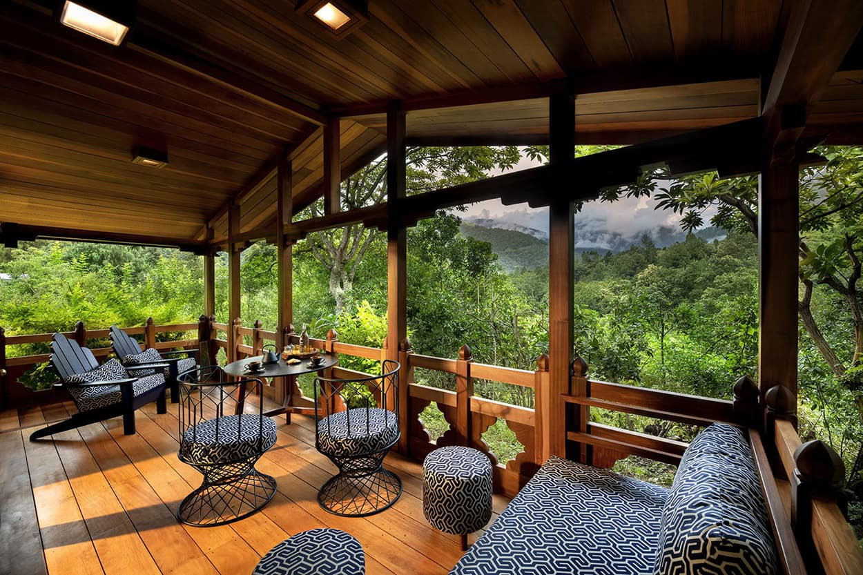 A cozy wooden balcony with a stunning view of lush greenery and mountains. It features comfortable seating with patterned cushions, a small table set for beverages, and a serene, natural ambiance.