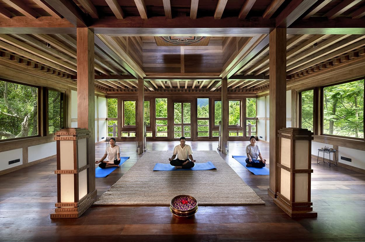 A serene meditation room with large windows overlooking greenery. Three individuals sit in a meditative posture on blue mats, surrounded by wooden beams and a light brown rug. A basket of flowers is placed in the foreground.