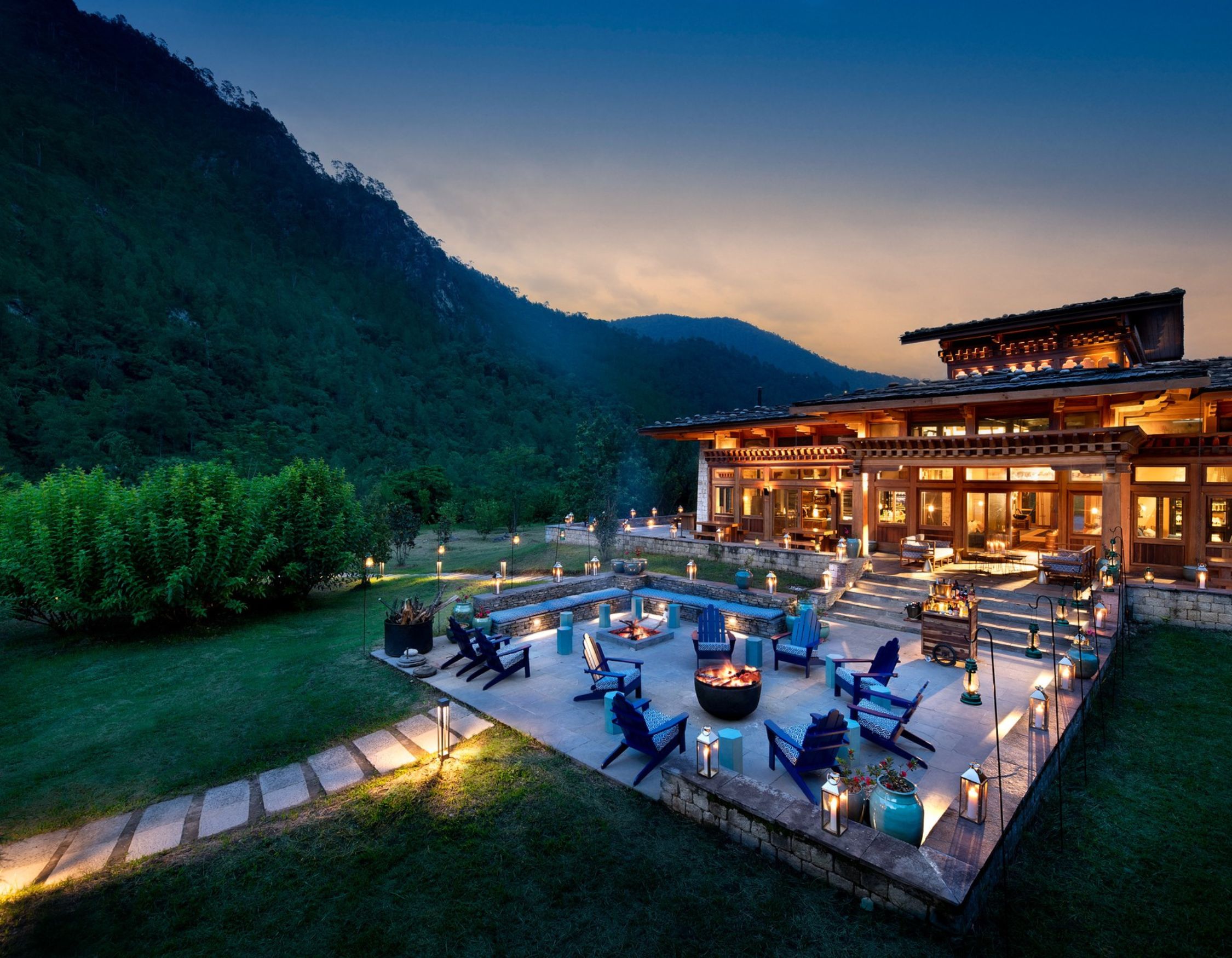 A beautiful mountain lodge at twilight, featuring a spacious patio with blue chairs and a fire pit surrounded by lanterns. Lush greenery and rocky hills create a serene backdrop. Warm lights illuminate the lodge's wooden architecture.