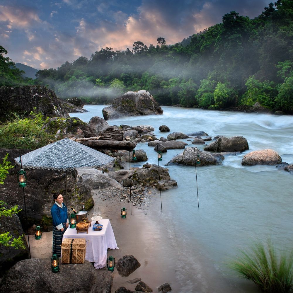 A serene riverside scene featuring a woman in traditional attire standing by a table set for a meal. The table is decorated with lanterns and surrounded by large rocks and lush greenery. Mist rises from the river under a dramatic sky.