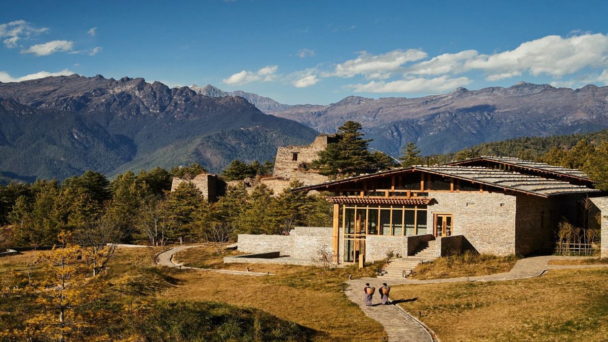 A modern building with large glass windows sits adjacent to ancient stone ruins, surrounded by lush greenery and mountain peaks under a clear blue sky. Two figures walk along a winding path toward the structure.