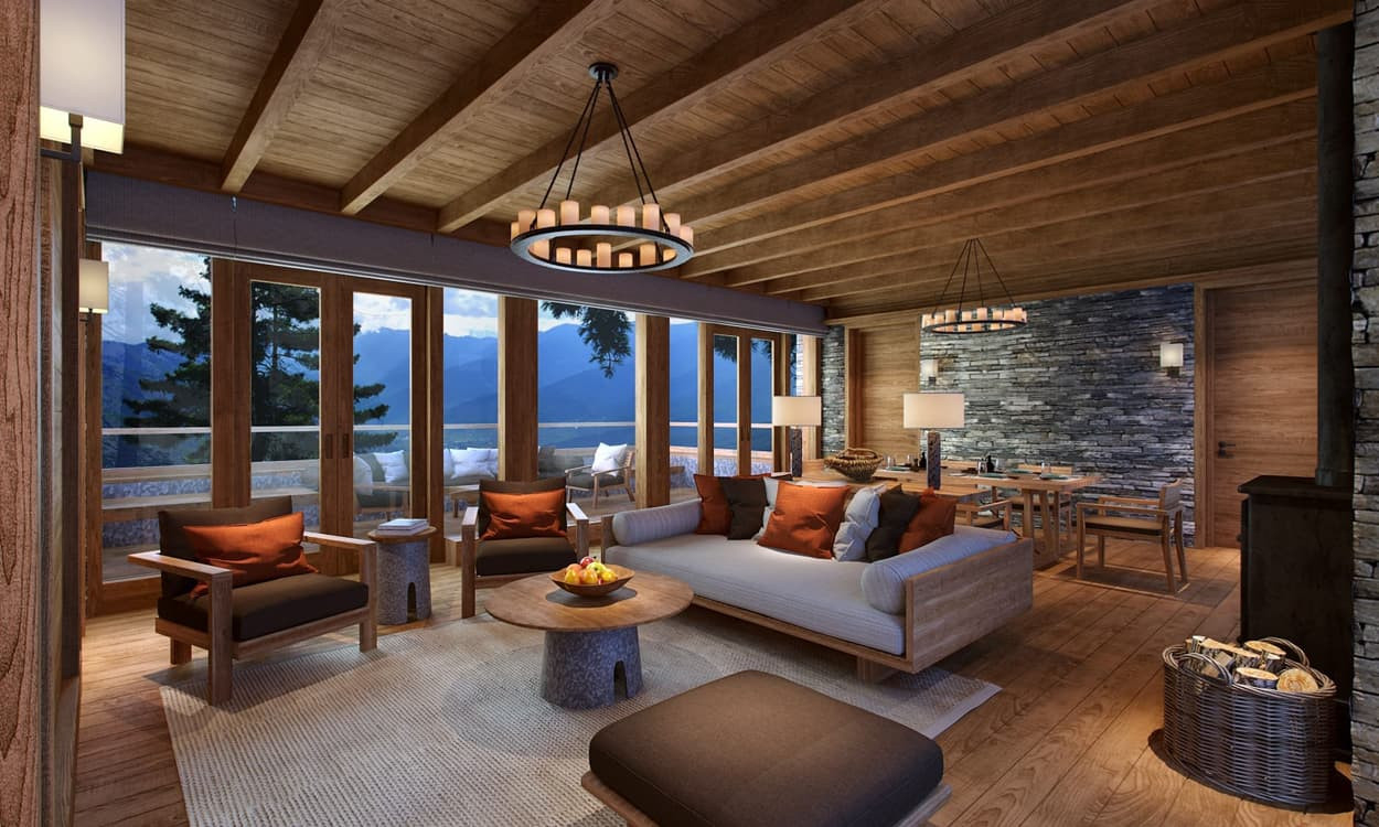 Cozy cabin interior with wooden ceilings and walls, featuring a spacious living area with a large sofa, accent chairs, and a coffee table. Large windows offer a scenic view of mountains and trees, while warm lighting creates an inviting atmosphere. A dining area with a wooden table is visible in the background.