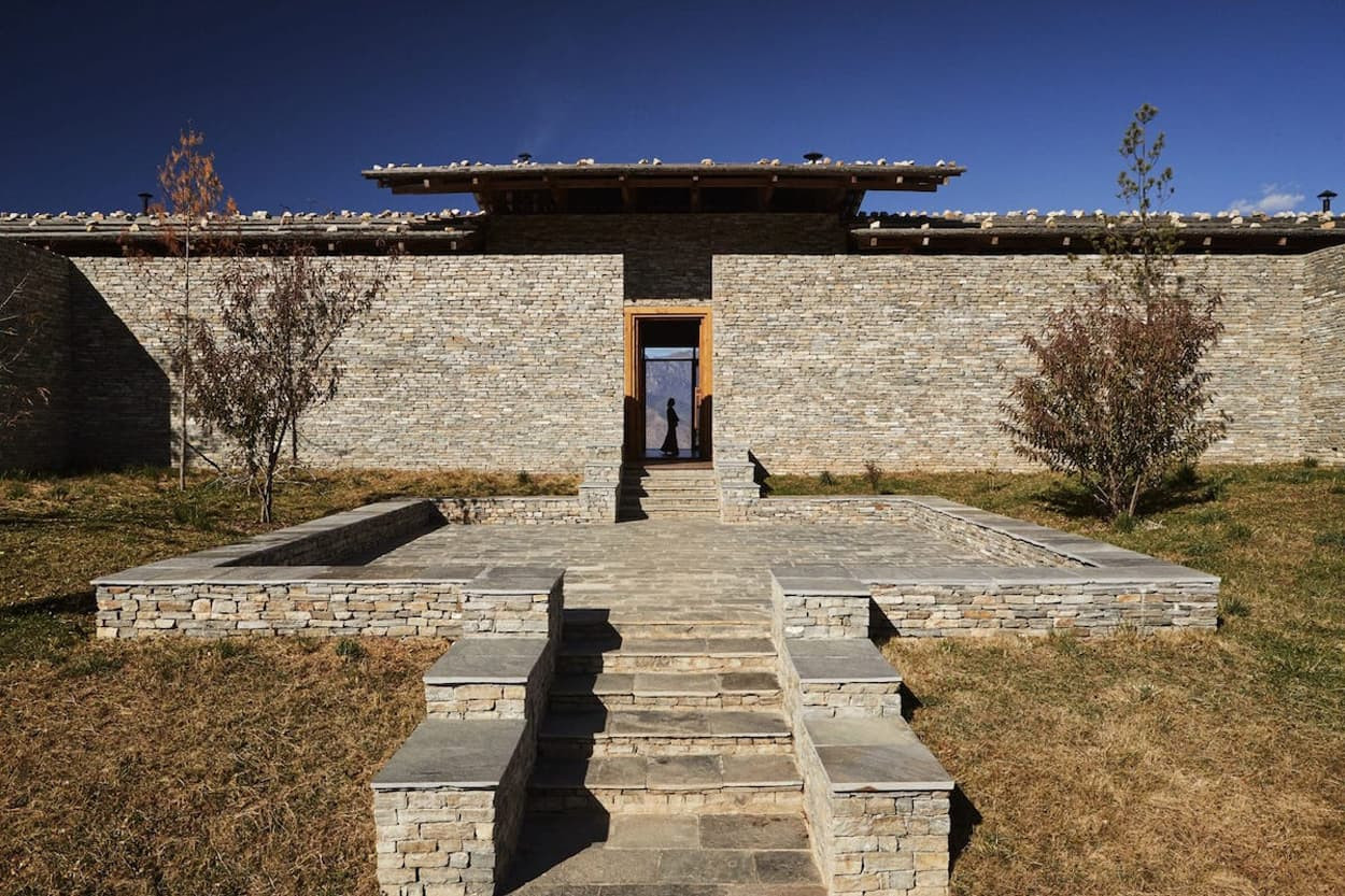 A modern stone building with a flat roof, flanked by trees and grass, features a pathway leading to a central entrance. The structure has a minimalist design, and a small courtyard area is visible in front of it.