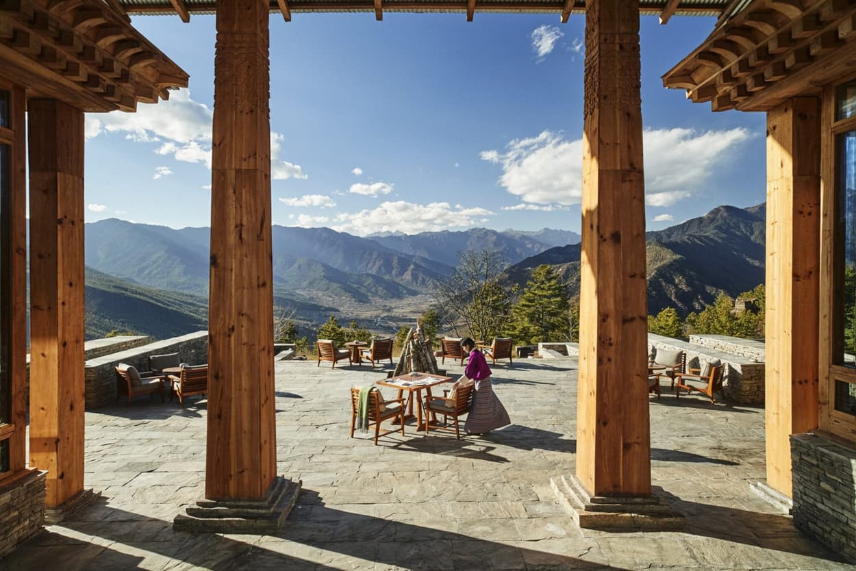 A scenic view from a stone terrace featuring wooden pillars, overlooking a valley with mountains in the distance. A woman in traditional attire stands near a table, surrounded by outdoor seating under a blue sky with fluffy clouds.