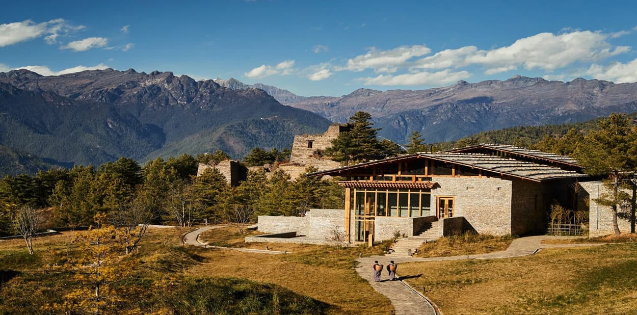 A modern building with large glass windows sits at the foot of a mountain range, surrounded by lush greenery and a winding path. In the foreground, two people walk towards the building, and ancient ruins are visible in the background under a clear blue sky with scattered clouds.