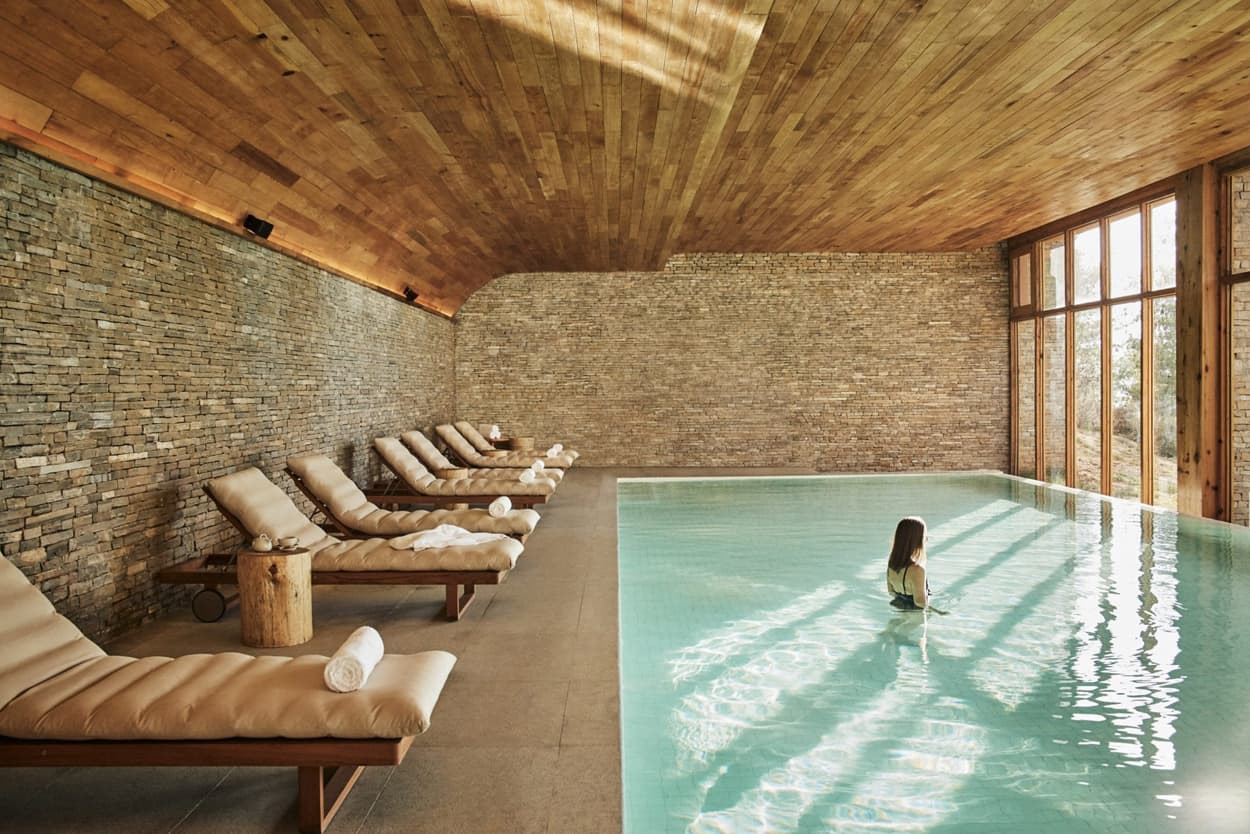 A serene indoor swimming pool features wooden beams and stone walls. Lounge chairs line one side, and a person is seen wading in the clear water, surrounded by natural light streaming in through large windows.