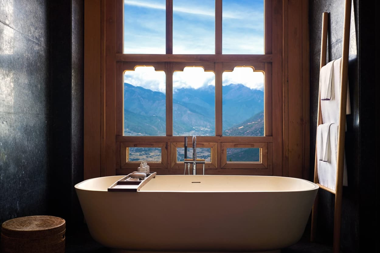 A modern bathtub sits in front of a large window, offering a stunning view of mountains and blue skies. The bathroom features dark stone walls, wooden accents, and neatly arranged towels hanging nearby.
