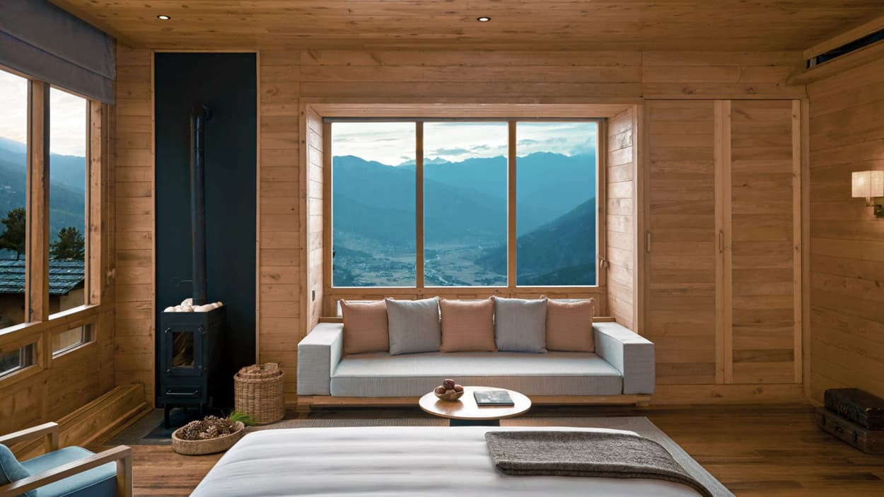 A cozy wooden room featuring a large window with a view of mountains. A comfortable sofa with cushions is positioned by the window, and a small round table is in front. A minimalist bed is visible, along with a decorative basket and a small stove. Natural light fills the space.