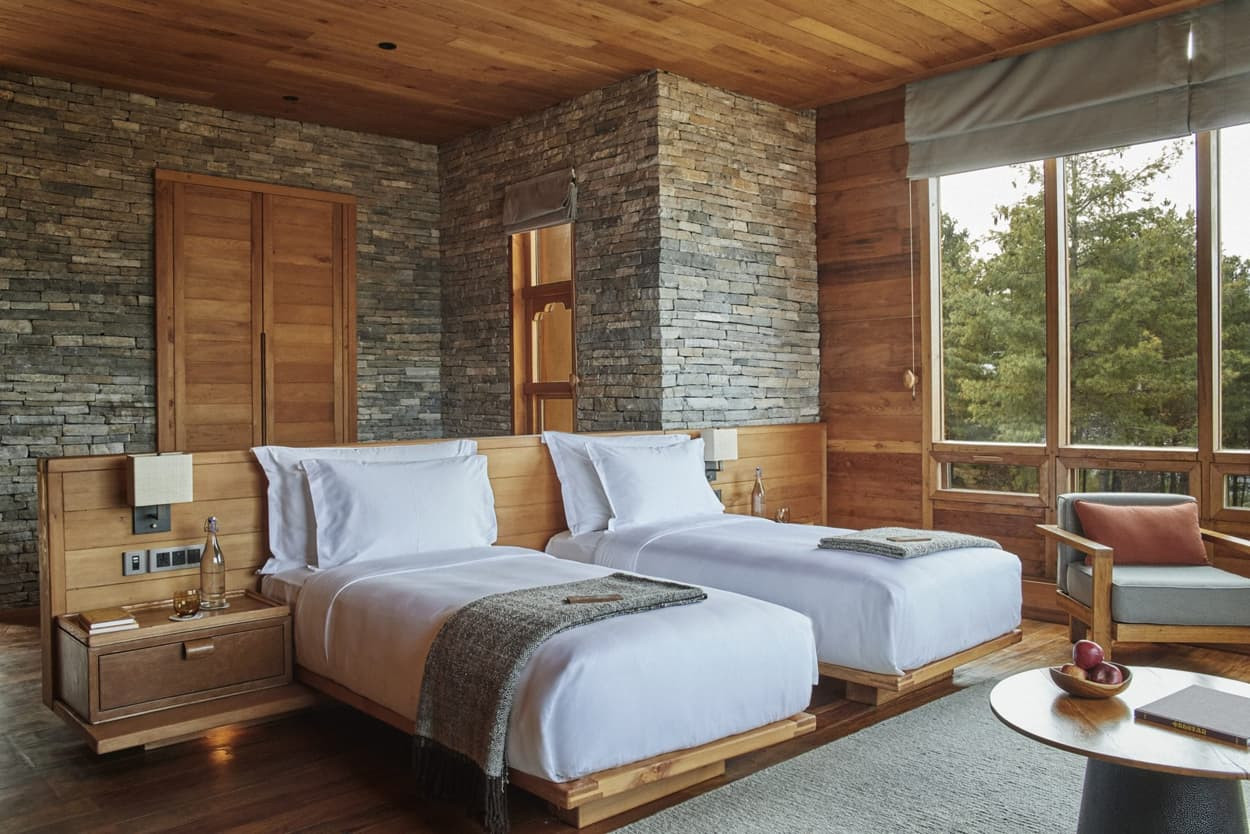 A cozy bedroom featuring two twin beds with white linens, a wooden headboard, and stone accent walls. Natural light floods the room through large windows, complemented by wooden flooring and a simple chair beside a small table with an apple.