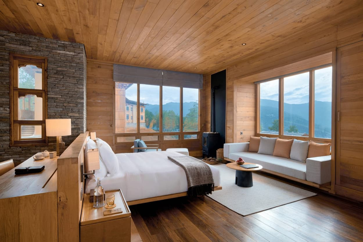 A spacious hotel room featuring wooden walls and floors, large windows with mountain views, a plush bed, a comfortable seating area, and minimalistic decor.