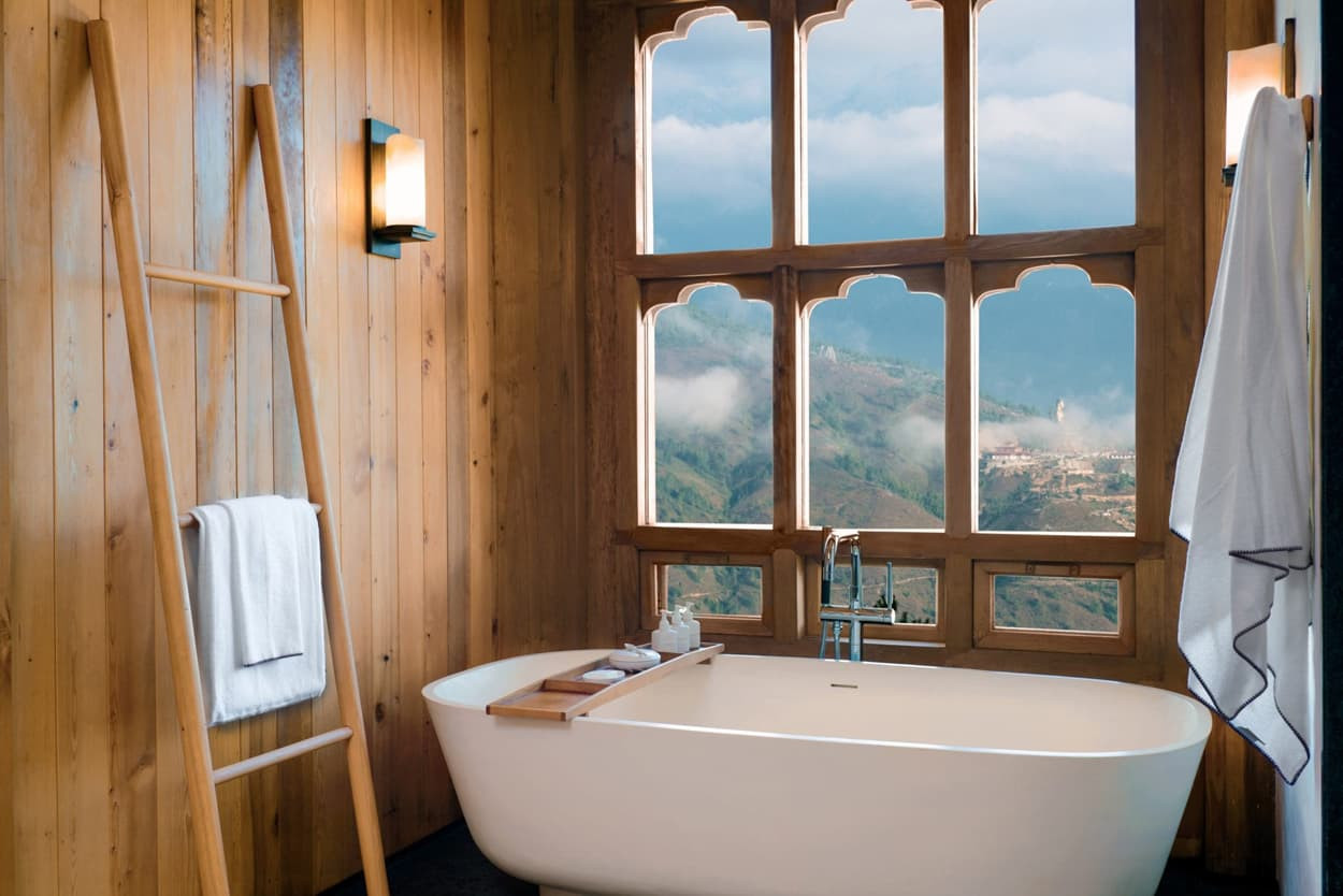 A modern, freestanding bathtub sits in a wooden bathroom. Large windows frame a scenic view of mountains and clouds outside. A towel hangs on a wooden ladder, and bath accessories are arranged on a wooden tray over the tub.