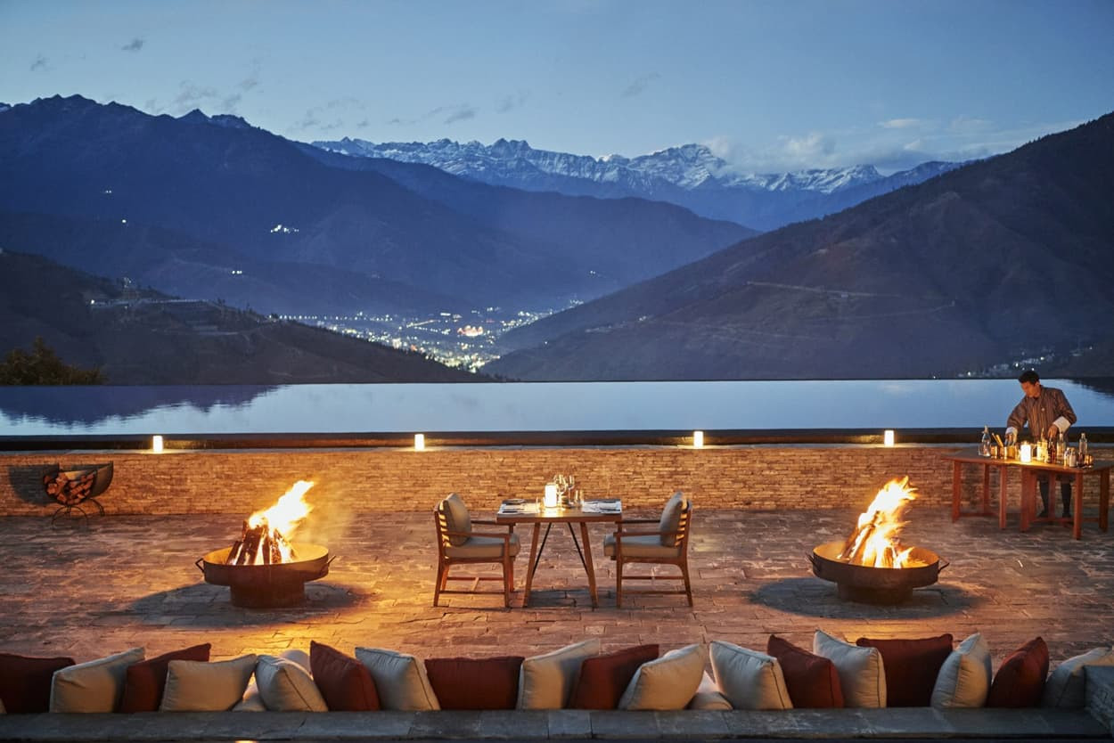 A cozy outdoor dining setup on a stone terrace, featuring a table with chairs surrounded by fire pits. In the background, mountains rise under a twilight sky, with distant lights twinkling in a valley below. A server is visible, setting up the dining area.