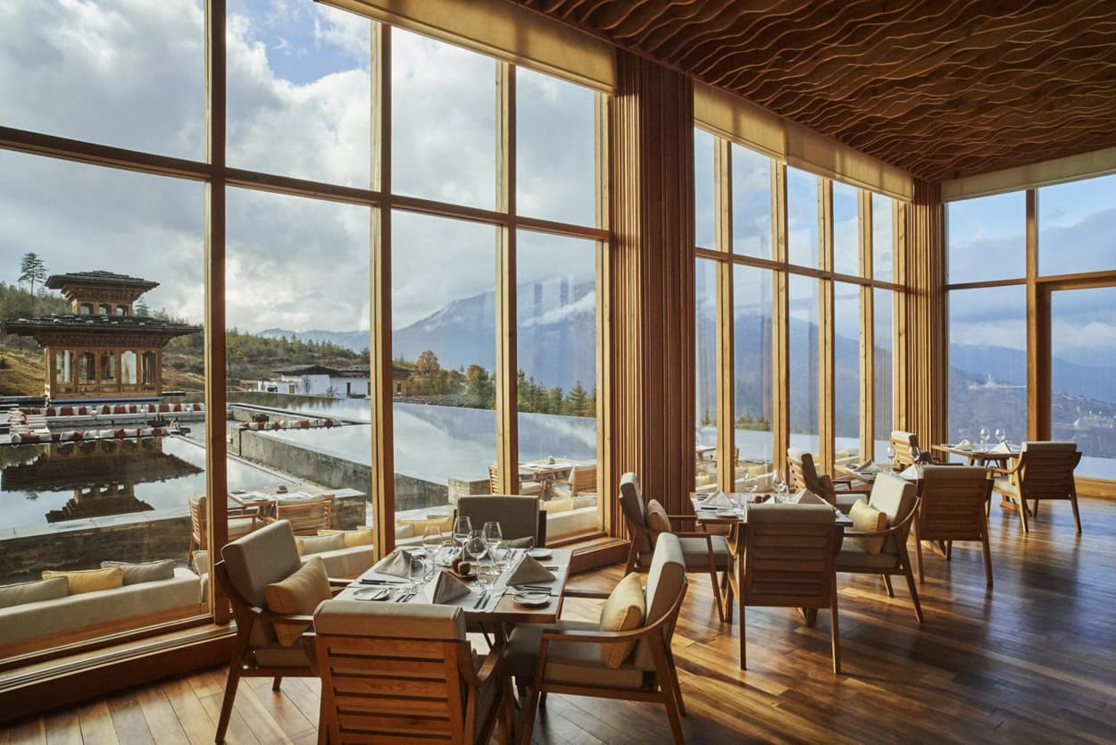 A spacious restaurant featuring large windows with stunning mountain views. Wooden interiors and elegant tables set for dining are visible, creating a serene and inviting atmosphere.