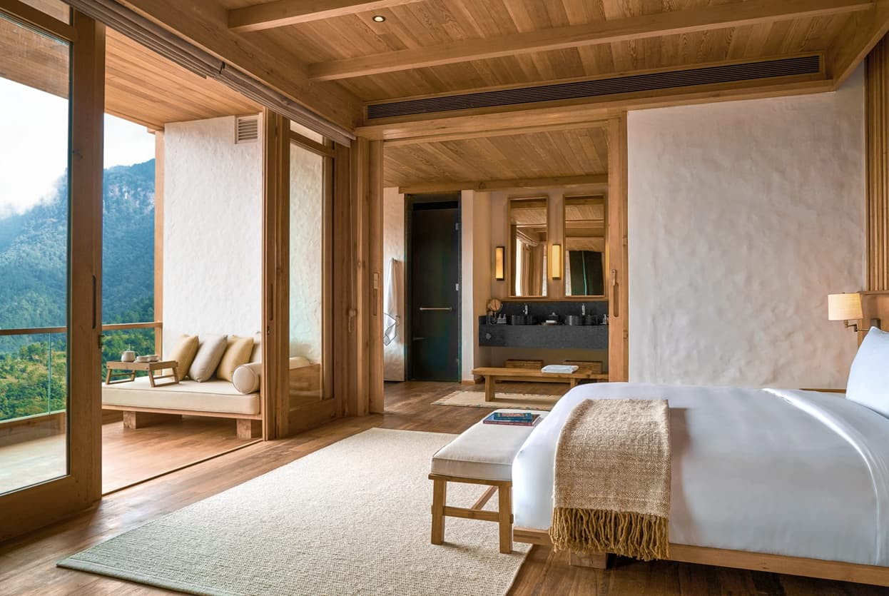 A modern, wooden interior of a luxurious hotel room featuring a large bed, a cozy seating area by expansive glass doors, and stunning views of the lush mountains outside. Natural light fills the space, highlighting the warm, earthy tones of the decor.