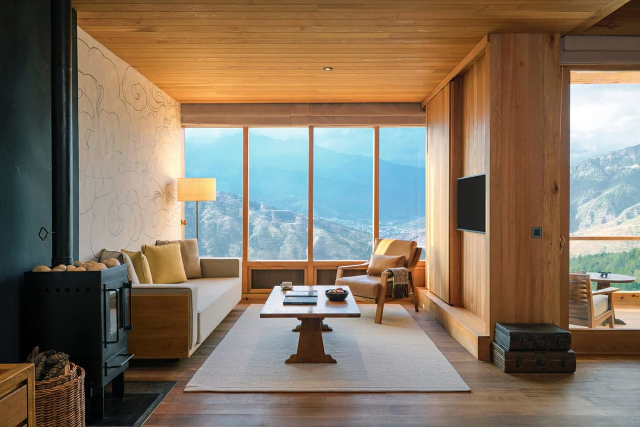 A modern living room with large windows showcasing a mountain view. The room features wooden walls and a light-coloured sofa, wooden coffee table, and a minimalist decor with a cozy atmosphere. A small stove adds warmth to the space.