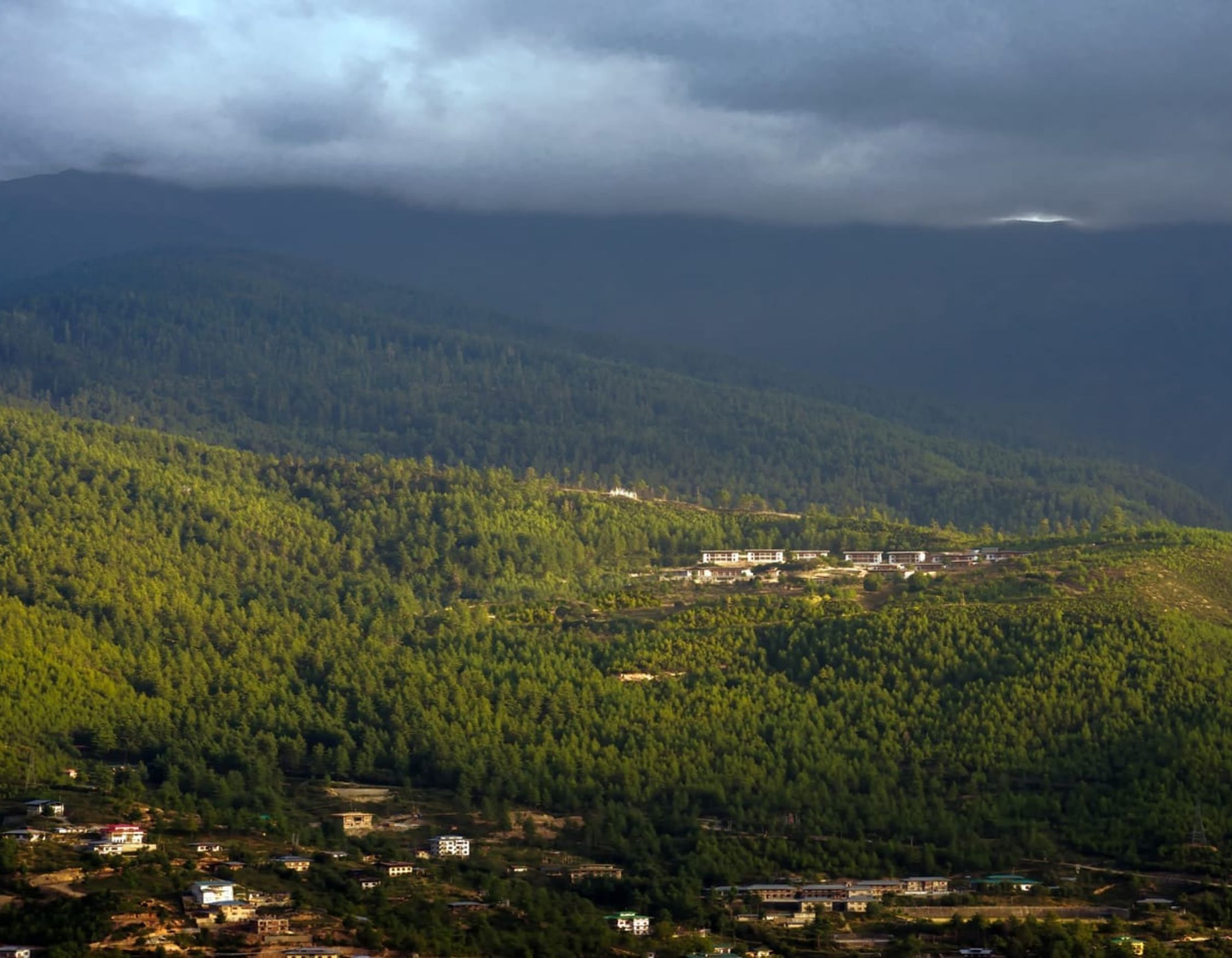 Lush green mountains with a mix of coniferous trees under a dramatic sky, displaying a blend of sunlight and shadow. In the background, a small village is nestled along the hillside.