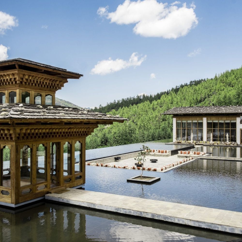 A traditional wooden structure with ornate details overlooks a serene, reflective pool surrounded by lush greenery and hills. In the background, a modern building features large windows and minimalist design. Blue skies with a few clouds enhance the tranquil atmosphere.