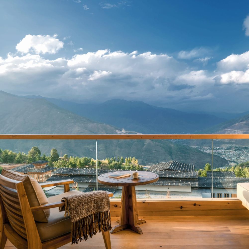 A cozy seating area features a wooden chair and round table on a balcony, offering a breathtaking view of mountains and cloudy skies in the background.