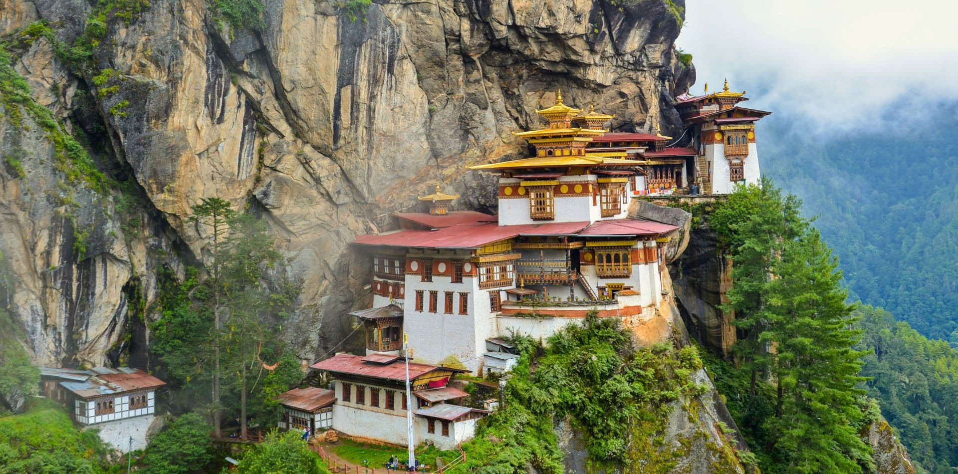 A stunning cliffside monastery with traditional Bhutanese architecture, surrounded by lush greenery and misty mountains. The building features golden roofs and multiple levels, perched dramatically on the edge of a rocky outcrop.