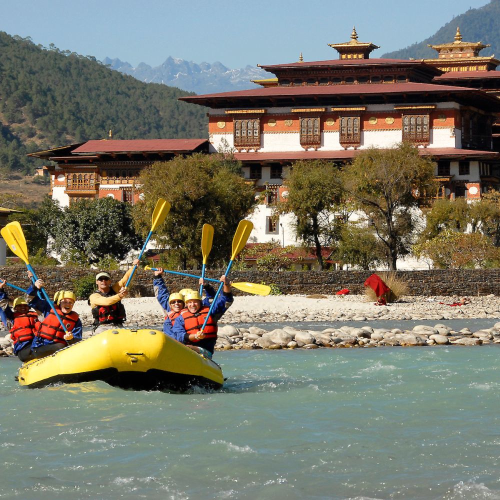 A group of four people in a yellow kayak paddle down a river, smiling and raising their arms. In the background, a traditional Bhutanese building with ornate architecture is surrounded by trees and mountains.