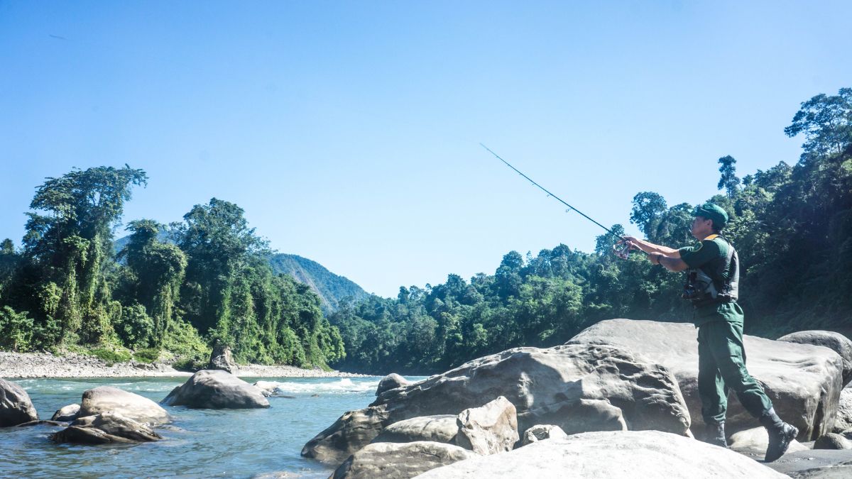 A person stands by a river, fishing with a rod. Lush green trees and mountains are visible in the background under a clear blue sky. Large rocks are scattered around the riverbank.