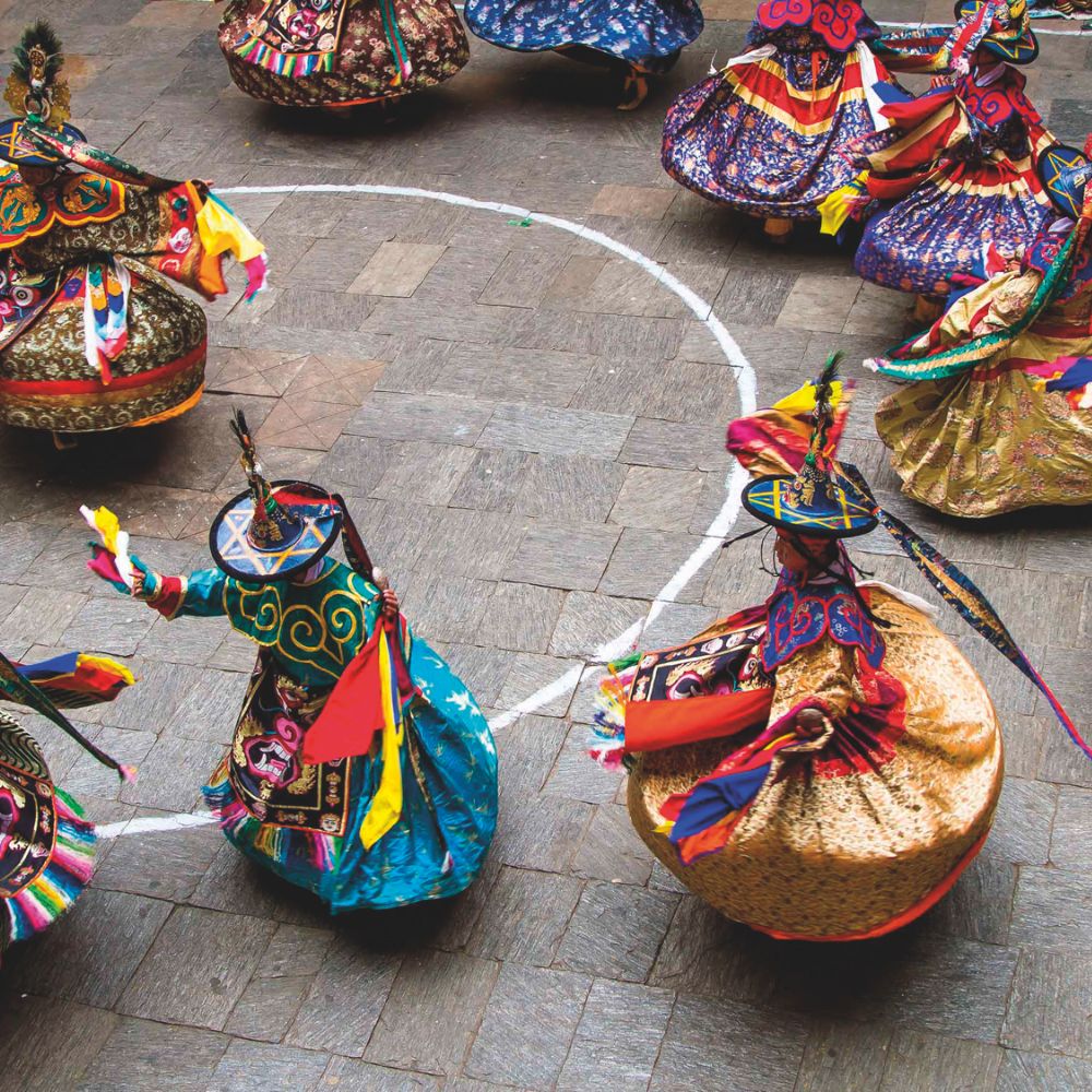 Colorfully dressed dancers perform in circular motion, showcasing vibrant traditional costumes adorned with intricate patterns and bright accessories. The dancers are arranged in a circular formation within a marked area on a stone floor.