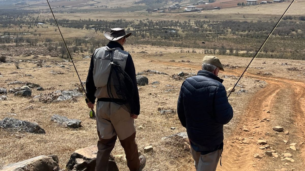 Two men walking in a rural landscape, carrying fishing rods. One man is wearing a hat and light clothing, while the other is in a dark jacket and cap. The terrain is dry with sparse vegetation and distant hills.