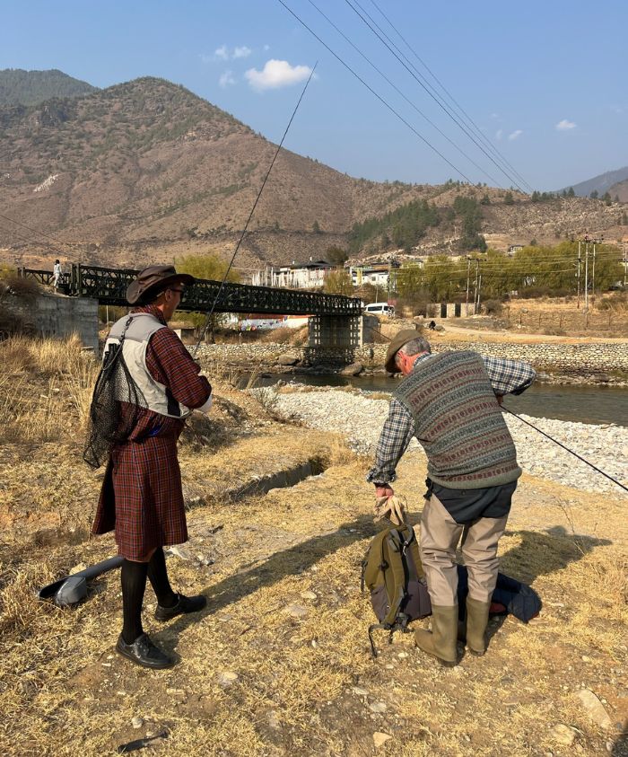 Two men stand by a river in a mountainous landscape. One man wears traditional clothing and stands with a fishing rod, while the other prepares gear. A bridge crosses the river in the background, surrounded by hills.