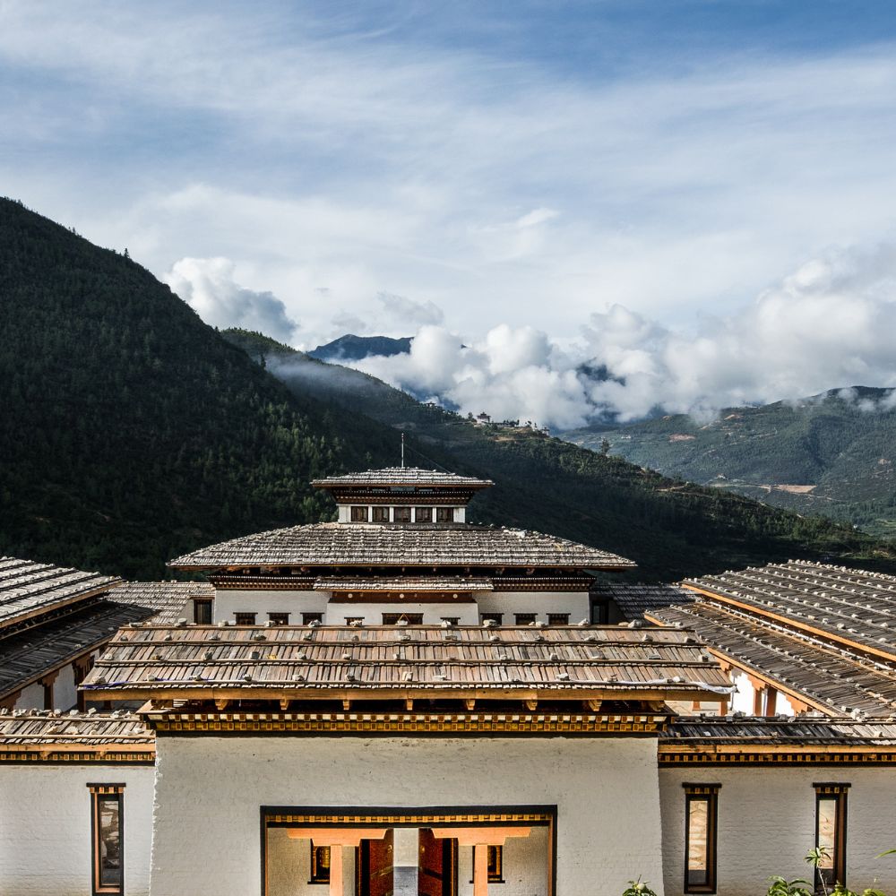 A view of a traditional Bhutanese building with a large, intricately designed roof, set against a backdrop of lush green mountains and a partly cloudy sky.