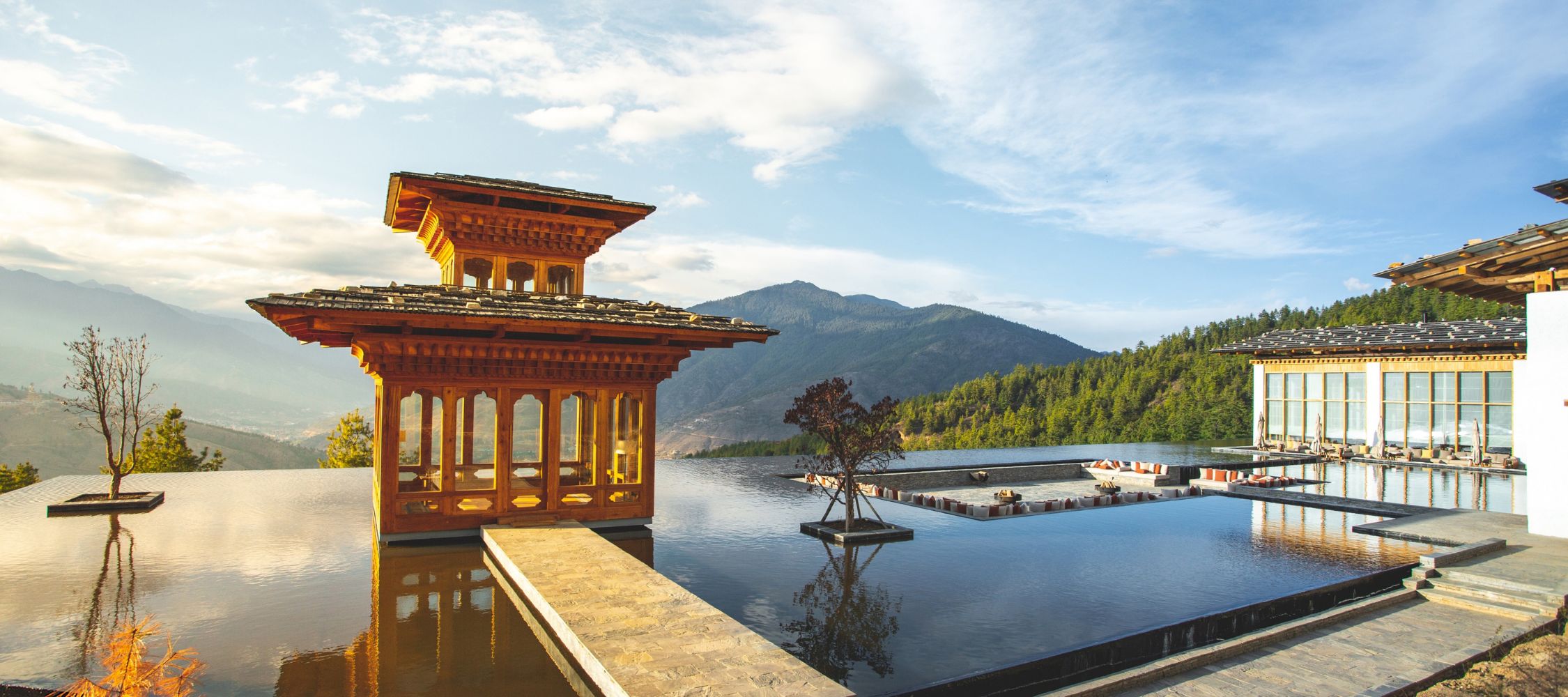 A stunning architectural structure with intricate wooden design stands over a reflective pool, surrounded by lush green mountains and a clear blue sky.