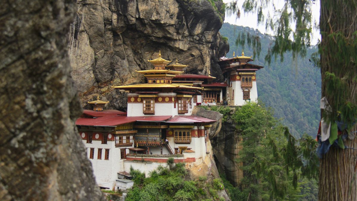 A cliffside monastery in Bhutan, featuring traditional architecture with ornate golden roofs and colorful decorations. The structure is surrounded by lush green mountains, creating a serene and picturesque scene.