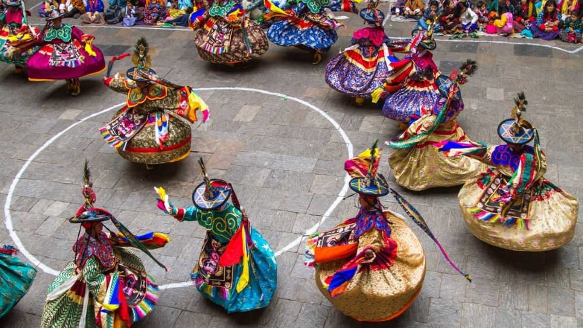 Vibrant dancers in traditional attire perform a lively dance, twirling in circular formation. Their costumes feature bright colours and intricate patterns, surrounded by an audience seated on the stone floor.