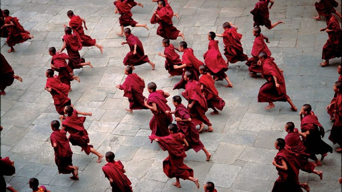 Monks in red robes run across a stone courtyard, creating dynamic movement as they participate in a group activity.