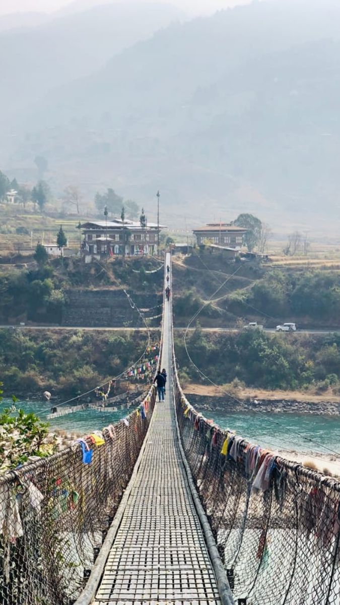A suspension bridge stretches across a river, connecting two sides of a valley. Colorful prayer flags decorate the sides, and a few people walk across the bridge. In the background, hills and a small house are visible, surrounded by misty mountains.