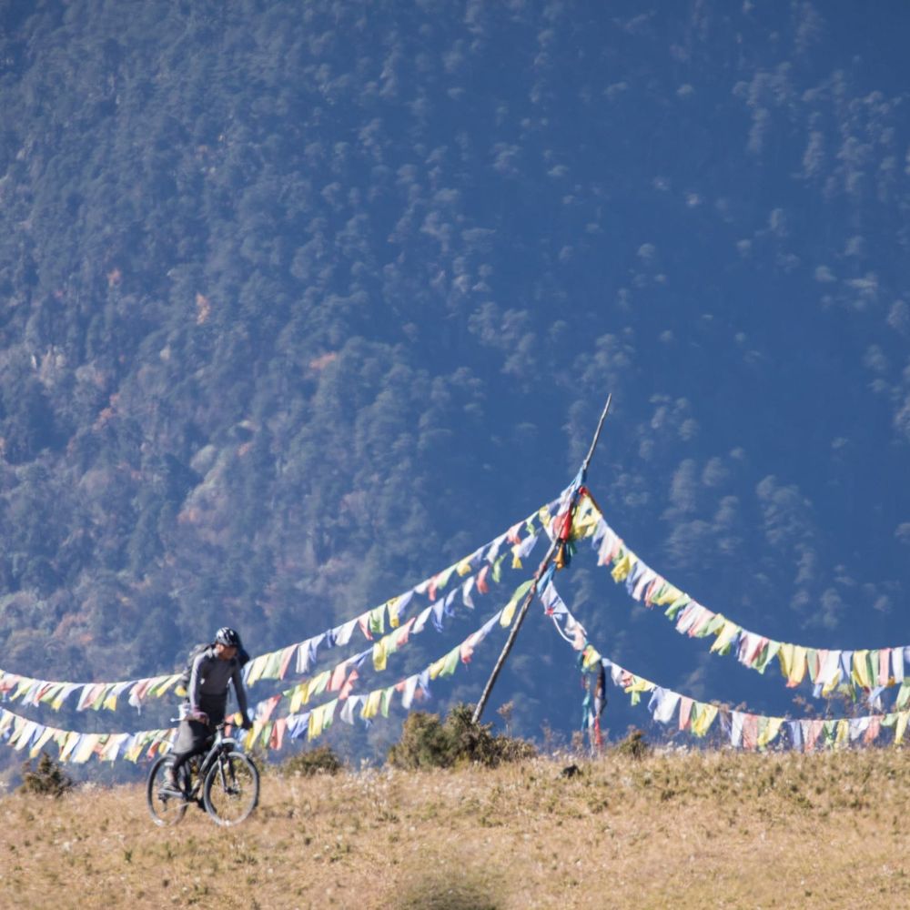 A cyclist rides along a grassy hill, with colorful prayer flags strung across the landscape against a backdrop of mountains and trees.