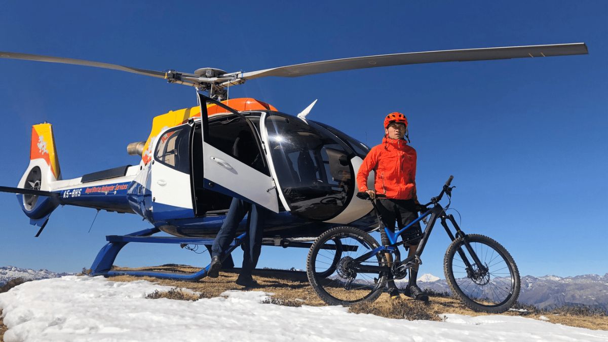 A mountain biker in an orange jacket and helmet stands beside a helicopter on a snowy mountain peak, with a bicycle positioned next to him. The landscape features snow-capped mountains under a clear blue sky.