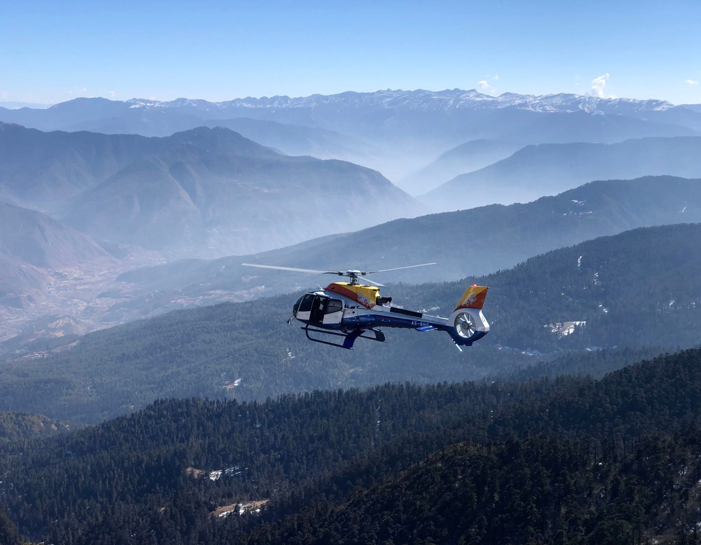 A helicopter flying above a mountainous landscape with snow-capped peaks in the distance. The terrain below features dense forests and valleys. Clear blue sky enhances the scenic view.
