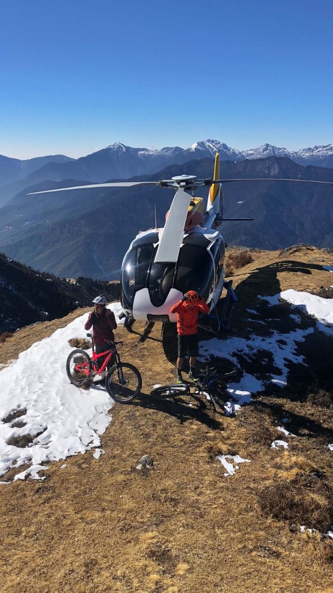 Two mountain bikers standing near a helicopter on a snowy mountain slope, with majestic peaks in the background. One biker is adjusting their bike while the other looks towards the helicopter. The sun shines brightly in a clear blue sky.