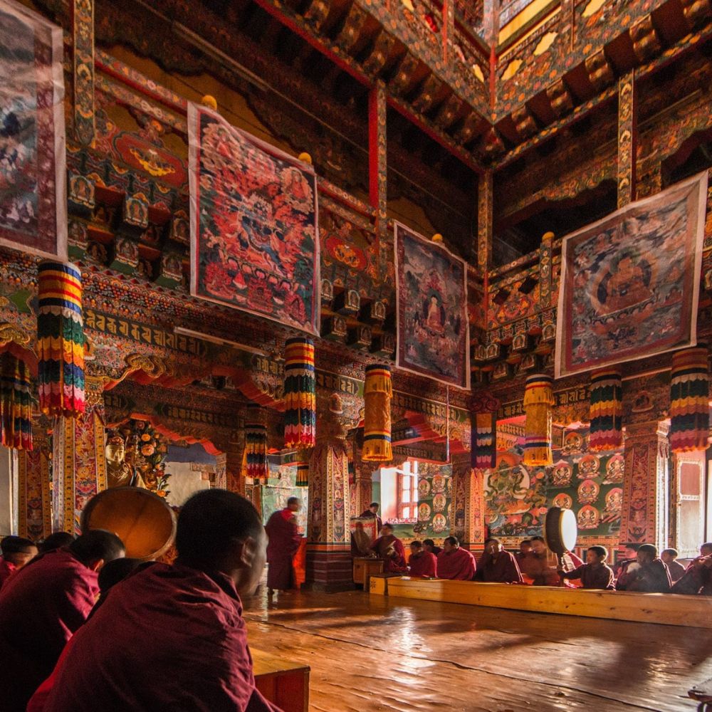 Monks in red robes sit on the floor of a vibrant, intricately decorated temple. Colourful wall hangings and paintings adorn the walls, while ornate pillars and hanging decorations add to the rich atmosphere. Sunlight filters through the windows, casting a warm glow.