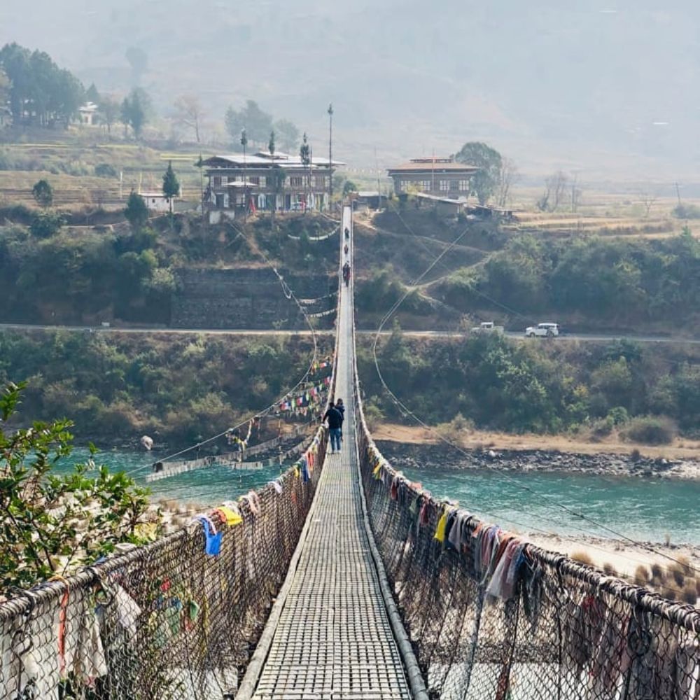 A long suspension bridge stretches over a river, with colorful prayer flags hanging along the sides. In the background, a few buildings and trees are visible, surrounded by a misty landscape.