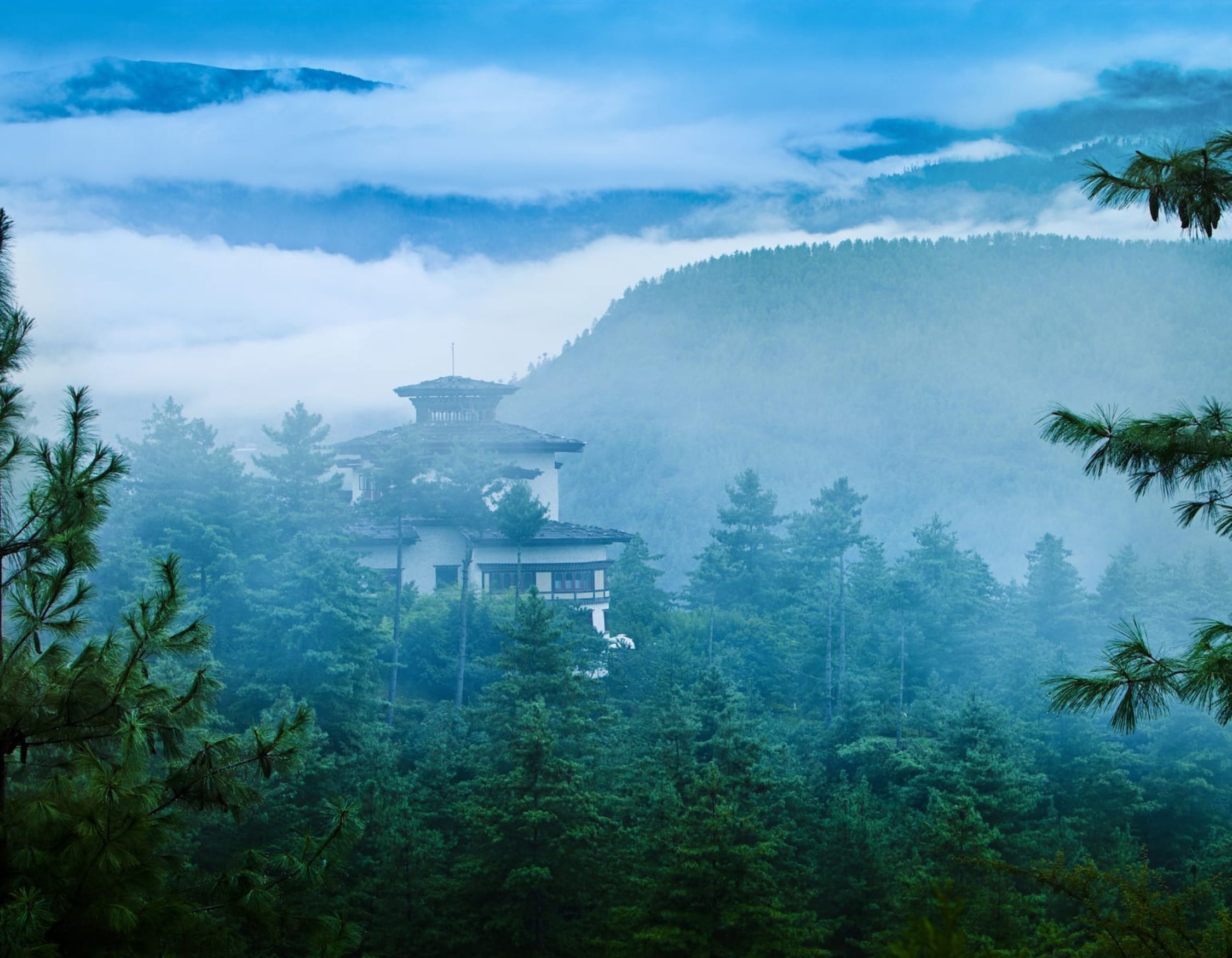 A serene landscape featuring a traditional building nestled among dense green trees, shrouded in mist with mountains in the background. The sky is a soft blue, adding to the tranquil atmosphere.